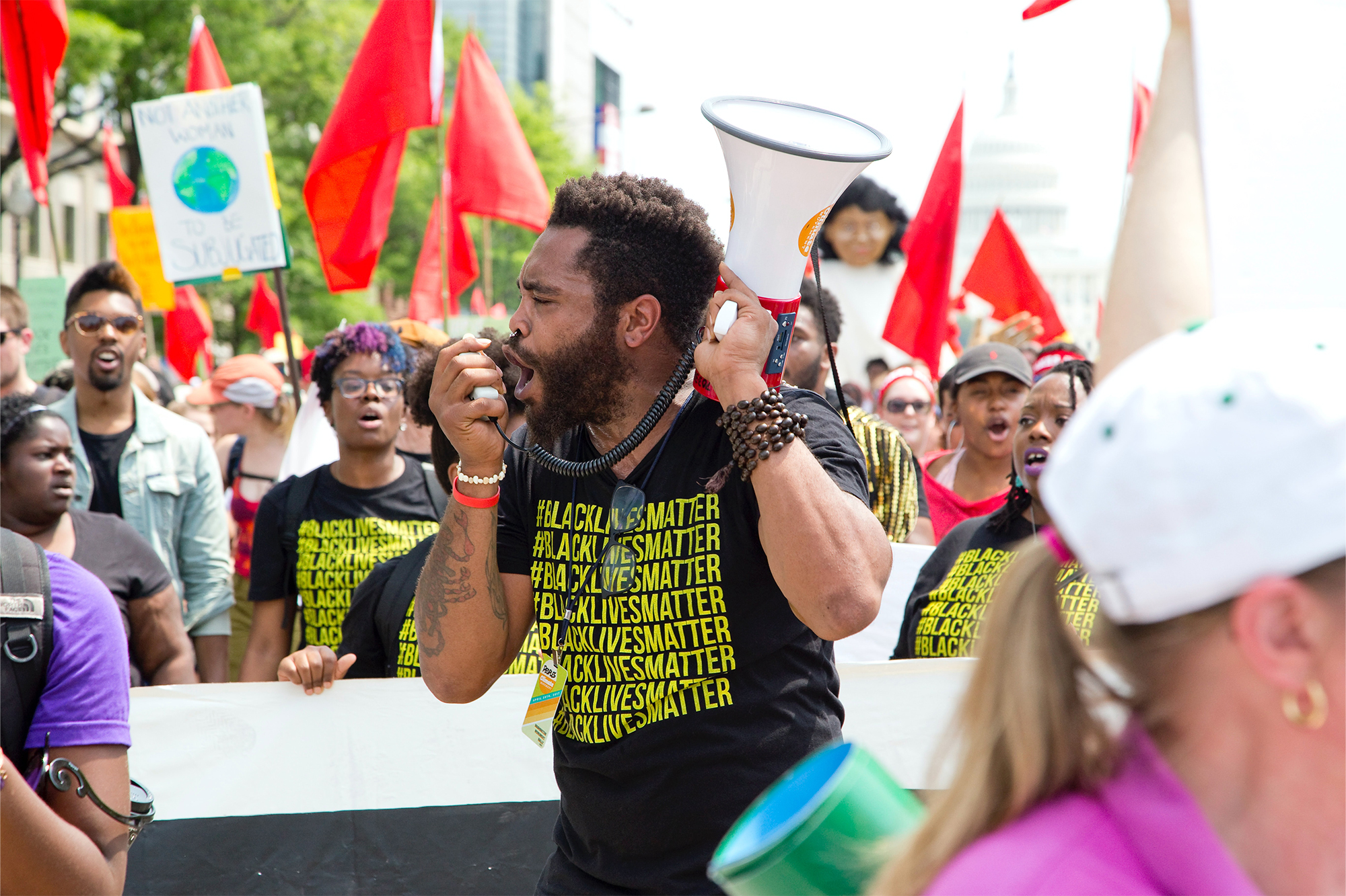 A man with a megaphone leads a Black Lives Matter protest, surrounded by others holding red flags and signs.
