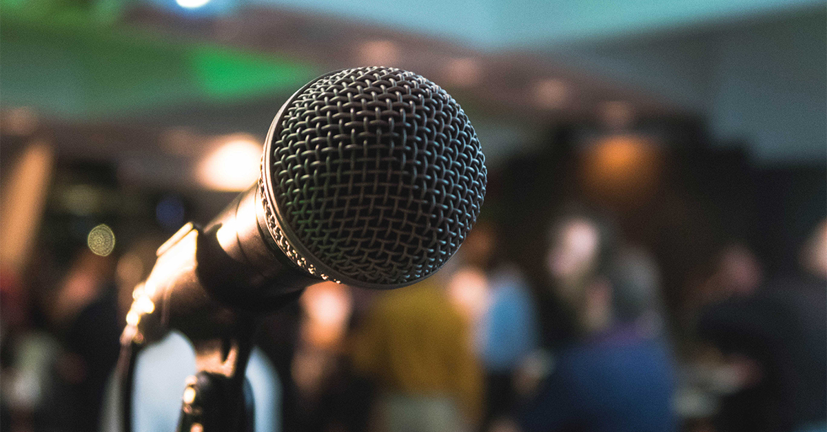 A close-up image of a microphone on a stand, with a blurred background suggesting a public event or gathering. The focus on the microphone implies that it is ready for someone to speak or perform, capturing the anticipation of a speech, presentation, or performance.